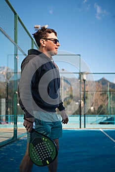 A man in a hoodie and shorts smiles while holding a padel tennis racket on a sunny outdoor court with mountains in the