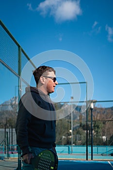 A man in a hoodie and shorts smiles while holding a padel tennis racket on a sunny outdoor court with mountains in the