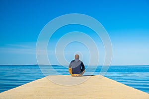 Man in hoodie sitting on the edge of dock and meditating. Sea horizon on the island od Pag, Adriatic sea, Croatia.