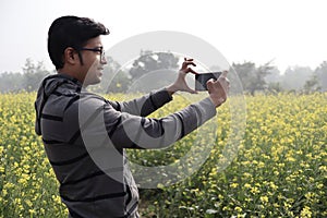 A man in hoodie and spectacles clicking nature photo with his smartphone in a mustard field with yellow blossomed
