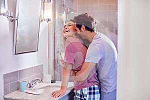 Man Hugging Woman Wearing Pyjamas In En Suite Bathroom As She Brushes Her Teeth