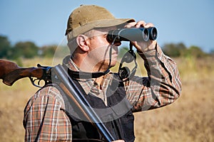 Man hunter with shotgun looking through binoculars in forest