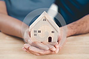 Man husband and Woman wife hands holding wooden house on the wood table background. family home, foster care, homeless charity