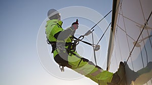 Man industrial climber in equipment and a helmet is tied with a rope to a building with a phone in his hands looking at