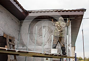 A man installs metal supports on the walls of the house for siding