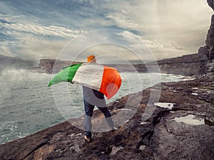Man with Irish flag standing on the edge of a cliff, stunning scenery with ocean, cliff and cloudy sky and fog over water in the