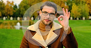 a man in a jacket and glasses looks at the camera in the park and shows ok