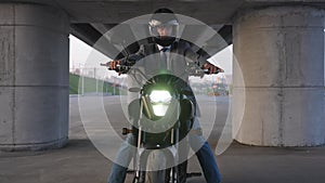 A man in a jacket and helmet sits on an electric motorcycle under a bridge. Retracting camera.
