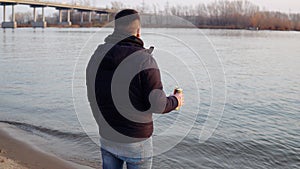 A man in a jacket stands on the riverbank, drinks a drink and throws a can into the river. Environmental pollution