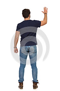 Man In Jeans And Blue T-shirt Is Standing With Arm Raised And Waving Hand. Rear View