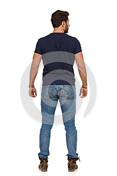 Man In Jeans And Blue T-shirt Is Standing Legs Apart And Looking Away. Rear View