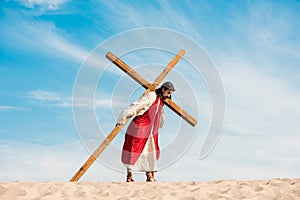 Man in jesus robe walking with wooden cross against sky in desert