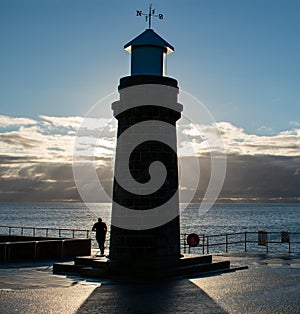 A man jogs around a lighthouse with him and the lighthouse is harp silhouette