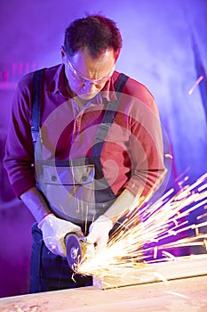 Man in jumpsuit goggles and gloves grinding metal with flow of sparks in garage