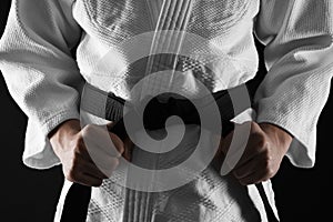 Man in keikogi tying black belt on dark background, closeup. Martial arts uniform