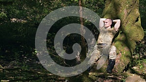 A man in a khaki uniform is resting in the forest under a tree