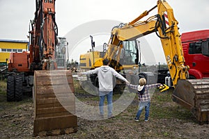 man and kid stand with their backs, look at construction site, where excavators with large buckets are located