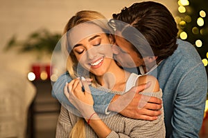 Man kissing neck of stunning woman, sitting near Christmas tree