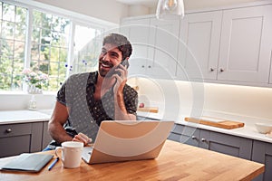 Man In Kitchen With Mobile Phone Working From Home Using Laptop During Health Pandemic