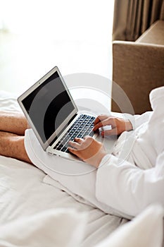 a man with a laptop on the bed. The man in the bedroom is lying on a bed with a computer on his lap and a notebook in