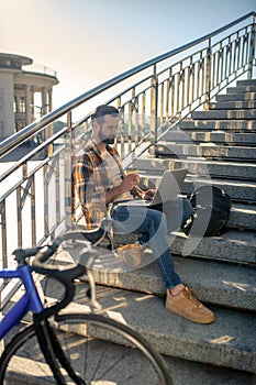 Man with laptop and coffee sitting on stairs near bike