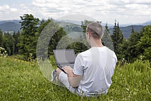 Man with laptop sitting on green grass on a background of mountains. Back view
