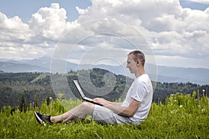 Man with laptop sitting on green grass on a background of mountains. Side view