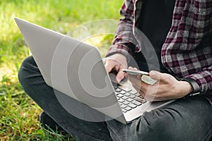 A man with a laptop and a smartphone sits on the grass, close-up.