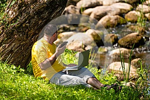man with laptop and smartphone working outside in the meadow by the river, outdoor office concept