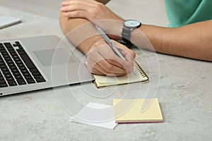 Man with laptop writing in notebook at table indoors, closeup