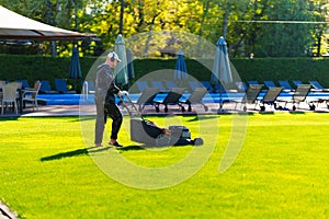 A man with a lawn mower mows grass on the lawn at a country club by the pool.