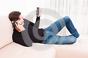 Man laying on sofa holding smartphone and remote control