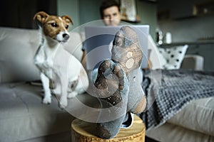 Man with leaky socks resting on sofa near dog, using laptop