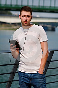 Man leaning on the fence using tablet and headphones, enjoying his surroundings by the river and old steel, green colored bridge.