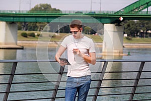 Man leaning on the fence using tablet and headphones, enjoying his surroundings by the river and old steel, green colored bridge.