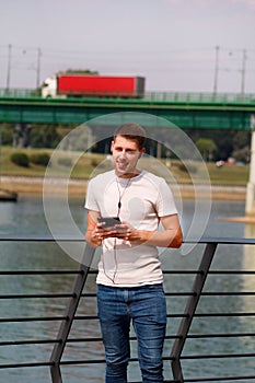 Man leaning on the fence using tablet and headphones, enjoying his surroundings by the river and old steel, green colored bridge.