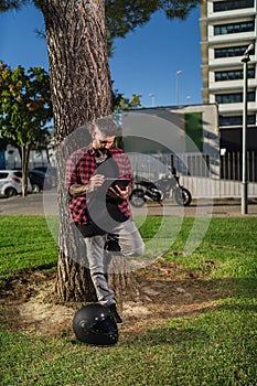 Man leaning on tree trunk, working on his tablet with a helmet next to him and a bike on background