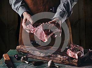 A man in a leather apron holds a piece of raw meat with a bone. Pork ribs in the hands of the butcher