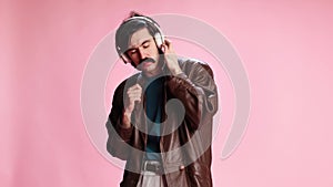 Man in leather jacket posing, listening to music in headphones and dancing over pink studio background. Concept of