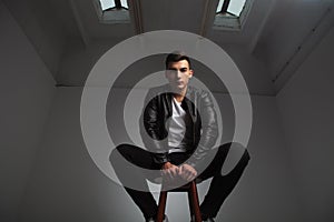 Man in leather jacket posing seated on stool in gray room