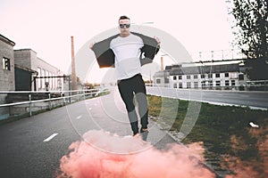 Man in a leather jacket standing at the street with smoke bomb. Colorful portrait of european teenager with red smoke bomb