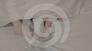 Man legs in white socks touched by two women with bare feet laying on bed on white bed sheets under blanket in close up