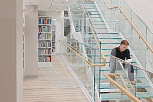 man in library is reading book sitting on steps of glass staircase