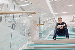 man in library is reading book sitting on steps of glass staircase