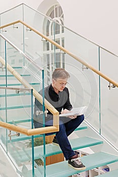 man in library is reading book sitting on steps of glass staircase