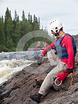 A man in a life jacket and helmet sits on a rock