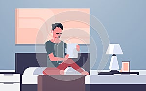 Man listening audio book through headphones guy using tablet sitting on bed modern home bedroom interior male character