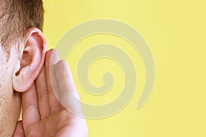 Man listens attentively with her palm to her ear, close up on yellow background, news concept