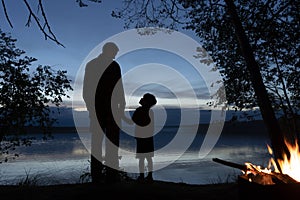 A man and a little boy father and son hold hands, stand on the shore and talk against the background of a bonfire and the reflecti