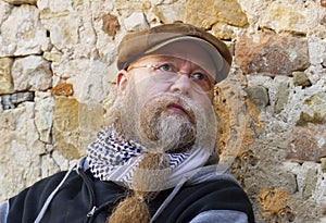 Man with long beard, glasses and cap, sits thoughtfully by a  wall